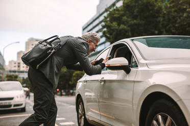 Mature businessman talking to taxi driver through opened window. Man taking a taxi for traveling in city. - JLPSF16389