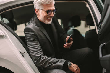 Smiling mature businessman sitting in the backseat of a taxi and looking at his phone. Senior male commuter taking a taxi ride in city. - JLPSF16396