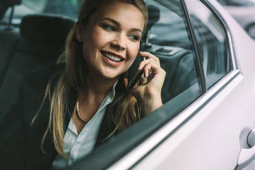 Lächelnde junge Geschäftsfrau, die auf dem Rücksitz eines Taxis sitzt und telefoniert. Weibliche Geschäftsfrau, die mit einem Taxi unterwegs ist und telefoniert. - JLPSF16405