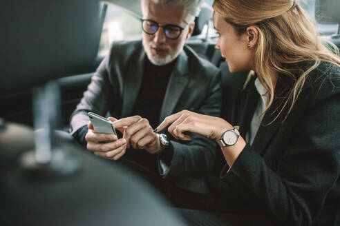 Junge Frau mit älterem Geschäftsmann, der ein Mobiltelefon benutzt, während er mit dem Auto unterwegs ist. Geschäftsleute, die ein Smartphone im Taxi benutzen. - JLPSF16408