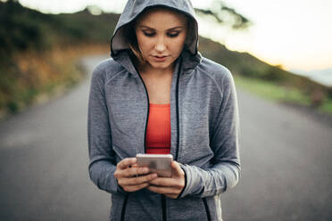 Fitness woman walking on street wearing a hooded sweat shirt looking at her mobile phone. Woman using her cell phone walking on an empty road. - JLPSF16449
