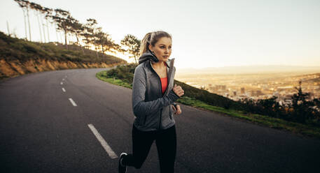 Frau in Fitnesskleidung läuft auf einer leeren Straße am Morgen. Fitness-Frau joggt auf einer hügeligen Straße. - JLPSF16452