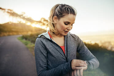 Läuferin, die eine Pause vom morgendlichen Joggen macht und auf ihre Armbanduhr schaut. Sportlerin, die ihre Armbanduhr auf einer hügeligen Straße am Morgen einstellt. - JLPSF16453