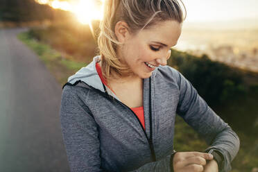 Smiling fitness woman setting a timer in her wrist watch standing outdoors. Female runner checking time in her watch during her morning run. - JLPSF16454