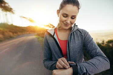 Lächelnde Fitness-Frau, die während ihres Morgenspaziergangs mit der Sonne im Hintergrund die Zeit überprüft. Porträt einer Läuferin, die beim morgendlichen Joggen ihre Armbanduhr einstellt. - JLPSF16458