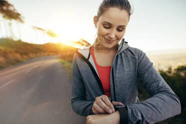 Smiling fitness woman checking time during her morning walk with sun in the background. Portrait of a female runner adjusting her wrist watch while jogging in the morning. - JLPSF16458