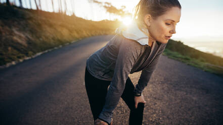 Female runner taking a break during her morning jog standing on a street with sun in the background. Woman athlete relaxing after workout resting her hands on her knees. - JLPSF16459
