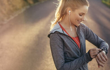 Fitness woman looking at her wrist watch during her morning fitness run. Smiling female athlete checking time standing on road while listening to music on earphones. - JLPSF16461