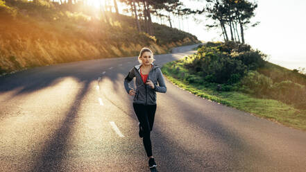 Fitness Frau läuft auf einer leeren Straße mit Sonne Flare im Hintergrund. Frau in Fitness-Kleidung läuft auf einer hügeligen Straße am Morgen. - JLPSF16462