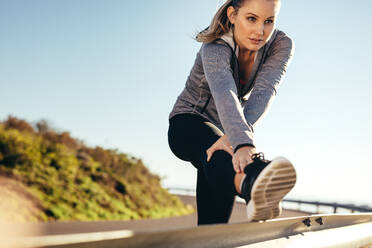 Eine Frau macht Dehnungsübungen auf dem Geländer einer Straße. Eine Frau in Fitnesskleidung beugt sich vor und streckt ihr Bein auf einem Geländer am Straßenrand. - JLPSF16464