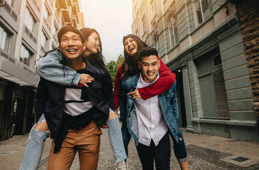 Zwei junge asiatische Männer tragen ihre Freundinnen auf dem Rücken auf einer Straße in der Stadt. Gruppe von Freunden, die im Freien auf einer Straße in der Stadt Huckepack nehmen. - JLPSF16483