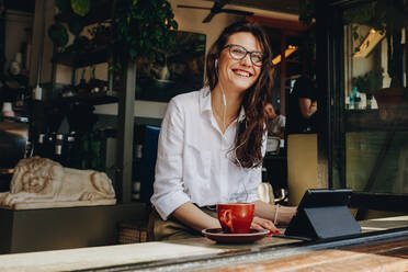 Businesswoman wearing earphones sitting at cafe table with digital tablet and cup of coffee, looking away smiling. Freelancer relaxing a coffee shop with digital tablet. - JLPSF16494