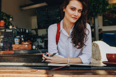 Beautiful woman sitting at coffee shop looking at camera. Confident female model wearing formal white shirt relaxing at cafe with her diary on table. - JLPSF16497