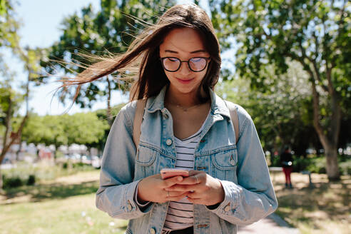 Porträt einer Studentin, die ein Handy benutzt, während sie in einem Park spazieren geht. Lächelndes junges Mädchen mit Brille und Jeansjacke, das ein Handy benutzt und im Freien steht. - JLPSF16512