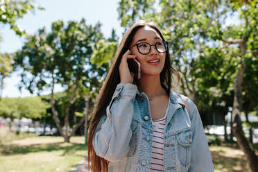 Asian girl in eyeglasses talking over mobile phone while waking in the street. Cheerful young girl talking over cell phone standing outdoors in a park. - JLPSF16513