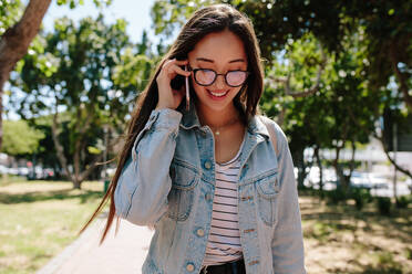 Nahaufnahme eines lächelnden College-Mädchens, das im Freien über ein Mobiltelefon spricht. Junges Mädchen mit Brille, das in einem Park spazieren geht und über ein Mobiltelefon spricht. - JLPSF16514