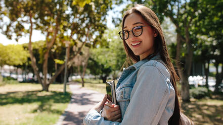 Side view of an asian college student standing on campus holding her books. Smiling girl in eyeglasses standing in a park on a sunny day. - JLPSF16516