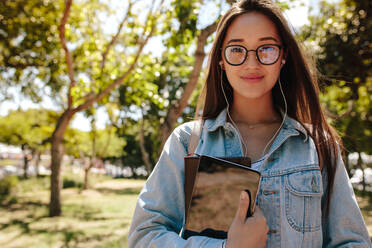 Nahaufnahme eines lächelnden asiatischen Mädchens mit Brille, das im Freien steht. Ein College-Student steht auf dem Campus und hält einen Tablet-PC und Bücher. - JLPSF16519