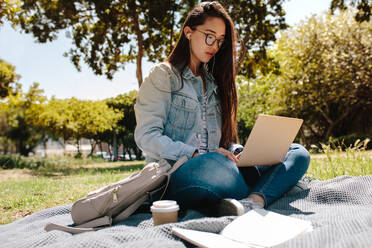 Nahaufnahme einer Studentin, die an einem sonnigen Tag auf einer Wiese sitzt und ihren Laptop benutzt. Asiatische Studentin, die an einem Laptop arbeitet und in einem Park sitzt und Musik hört. - JLPSF16520