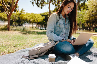 Lächelnd asiatischen College-Mädchen mit einem Laptop-Computer sitzen in der College-Campus an einem sonnigen Tag. Junges Mädchen tun ihre College-Arbeit auf Laptop sitzen im Freien, während Musik hören. - JLPSF16521