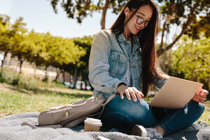 Nahaufnahme einer Studentin mit Brille, die in einem Park sitzt und ihren Laptop benutzt. Lächelndes asiatisches Mädchen, das im Freien sitzend am Laptop arbeitet, während es Musik hört. - JLPSF16522