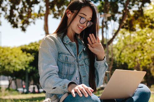 Nahaufnahme eines College-Mädchens bei einem Videogespräch in einem Park. Asiatisches Mädchen bei einem Videochat mit einem Laptop und Kopfhörern im Freien. - JLPSF16523