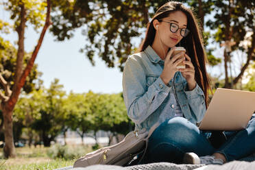 Fröhliches asiatisches College-Mädchen, das in einem Park sitzt und einen Laptop benutzt, während es einen Kaffee genießt. Nahaufnahme eines lächelnden Mädchens, das draußen in einem Park sitzt und sich mit einer Tasse Kaffee und Musik entspannt. - JLPSF16524