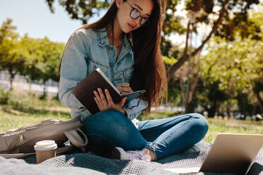 Nahaufnahme eines asiatischen Mädchens, das sich Notizen in ihrem Tagebuch macht und auf einen Laptop schaut. Mädchen mit Brille sitzt im Freien auf einer Wiese und studiert. - JLPSF16525
