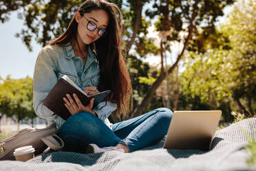 Nahaufnahme einer Studentin, die in einem Park sitzt und in ihr Notizbuch schreibt. Eine Studentin macht sich Notizen und schaut auf ihren Laptop. - JLPSF16526