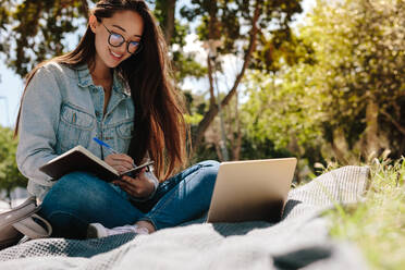 Nahaufnahme eines fröhlichen Mädchens, das in einem Park sitzt und lernt. College-Mädchen macht sich Notizen in einem Buch mit einem Laptop vor sich. - JLPSF16527