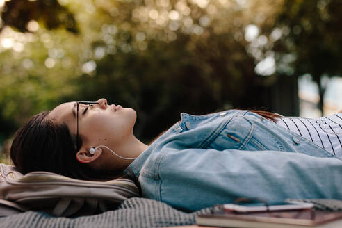 Nahaufnahme einer Frau, die in einem Park schläft und Musik hört. Ebenerdige Aufnahme einer jungen Frau, die mit einer Brille auf dem Boden liegt und Bücher neben sich hat. - JLPSF16529