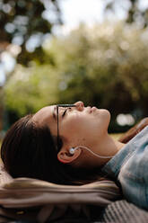 Nahaufnahme einer jungen Frau, die in einem Park auf dem Boden liegt und eine Sonnenbrille trägt. Frau entspannt sich im Freien und hört Musik. - JLPSF16530