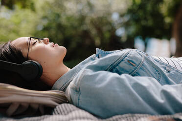 Seitenansicht einer Frau, die sich im Freien entspannt und mit Kopfhörern Musik hört. Nahaufnahme einer jungen Frau, die im Freien in einem Park schläft und die milde Sonne genießt und sich entspannt. - JLPSF16531