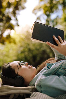 Nahaufnahme einer jungen Frau, die auf dem Boden liegt und mit Kopfhörern ein Buch liest. Frau in entspannter Stimmung, die im Freien liegend ein Buch liest und dabei Musik hört. - JLPSF16532