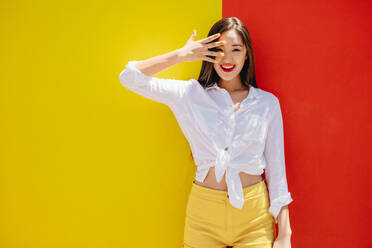 Smiling asian girl standing against a colorful background. Cheerful girl in colorful clothes standing outdoors with her hand to her face. - JLPSF16533