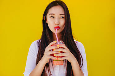 Portrait of an asian woman drinking juice using a straw. Young woman drinking juice in a disposable glass looking away. - JLPSF16537