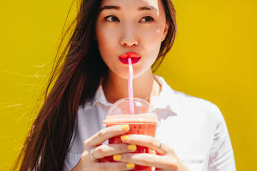 Asian woman drinking juice in a disposable plastic glass using a straw. Portrait of a young woman enjoying a glass of juice. - JLPSF16538