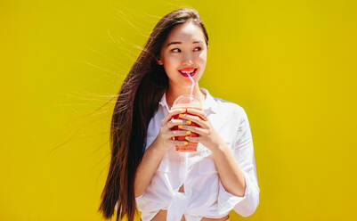 Smiling asian woman drinking juice in a disposable glass standing outdoors. Cheerful young woman holding a glass of juice and looking away. - JLPSF16539