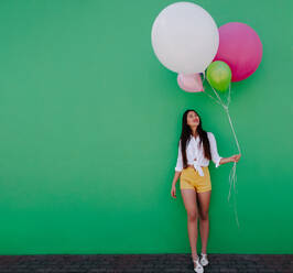 Smiling woman walking holding a bunch of balloons. Cheerful asian woman looking at a bunch of colorful balloons she is holding. - JLPSF16545