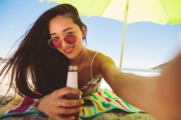 Nahaufnahme einer Frau, die am Strand unter einem Sonnenschirm liegt und ein Getränk hält. Lächelnde asiatische Frau im Urlaub am Strand an einem sonnigen Tag. - JLPSF16549
