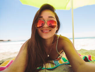 Porträt einer lächelnden Frau, die an einem sonnigen Tag am Strand im Schatten eines Sonnenschirms liegt. Nahaufnahme einer lächelnden Frau mit Sonnenbrille, die sich am Strand entspannt. - JLPSF16552