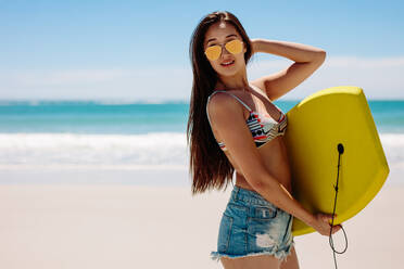 Side view of a woman carrying a bodyboard enjoying her vacation at the beach. Portrait of a woman in sunglasses at the beach holding a bodyboard. - JLPSF16556
