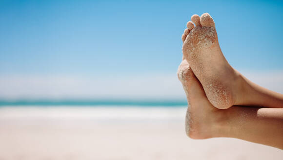 Nahaufnahme der Füße einer Person, die sich an einem ruhigen Strand an einem sonnigen Tag entspannt. Beine einer Person am Strand in einer entspannten Position mit einem Bein über dem anderen. - JLPSF16562