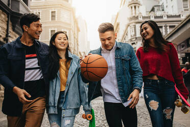 Eine Gruppe von Freunden, die gemeinsam auf der Straße spazieren gehen, wobei die Frauen ein Skateboard halten und der Mann einen Basketball wirft. - JLPSF16569