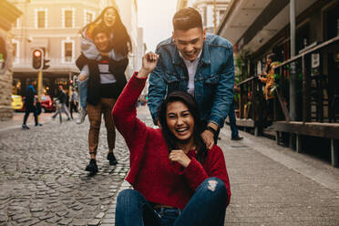 Excited woman being pushed on skateboard by man outdoors on street, with friends piggybacking in background on the city street. Group of friends hanging out outdoors in city. - JLPSF16579