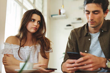 Frau sieht unglücklich aus, während ihr Mann ihr keine Aufmerksamkeit schenkt und mit seinem Handy beschäftigt ist. Schmollende Frau sitzt neben einem Mann, der während eines Dates Textnachrichten liest. - JLPSF16582