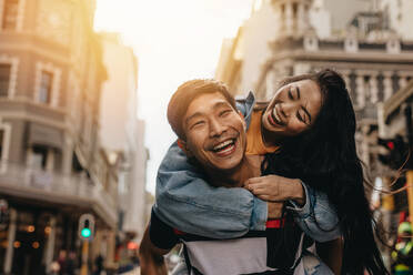 Asiatischer Mann, der seine Freundin im Freien auf der Straße huckepack nimmt. Fröhliches Paar, das sich in der Stadt amüsiert. - JLPSF16589