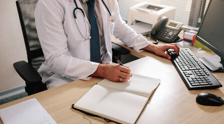 Hands of male doctor searching new information on medication using desktop computer at his desk. Medical professional using computer and writing notes in diary. - JLPSF16609