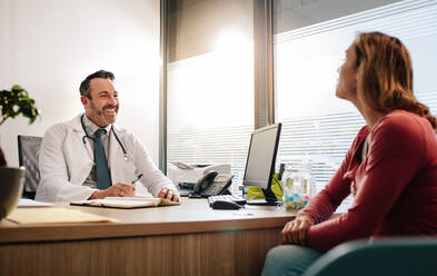 Doctor talking with female patient sitting across the desk in clinic. Mature male doctor in white medical coat consulting his patient. - JLPSF16612