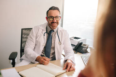 Male doctor sitting at his desk consulting a patient. General practitioner talking with patient in his clinic. - JLPSF16613
